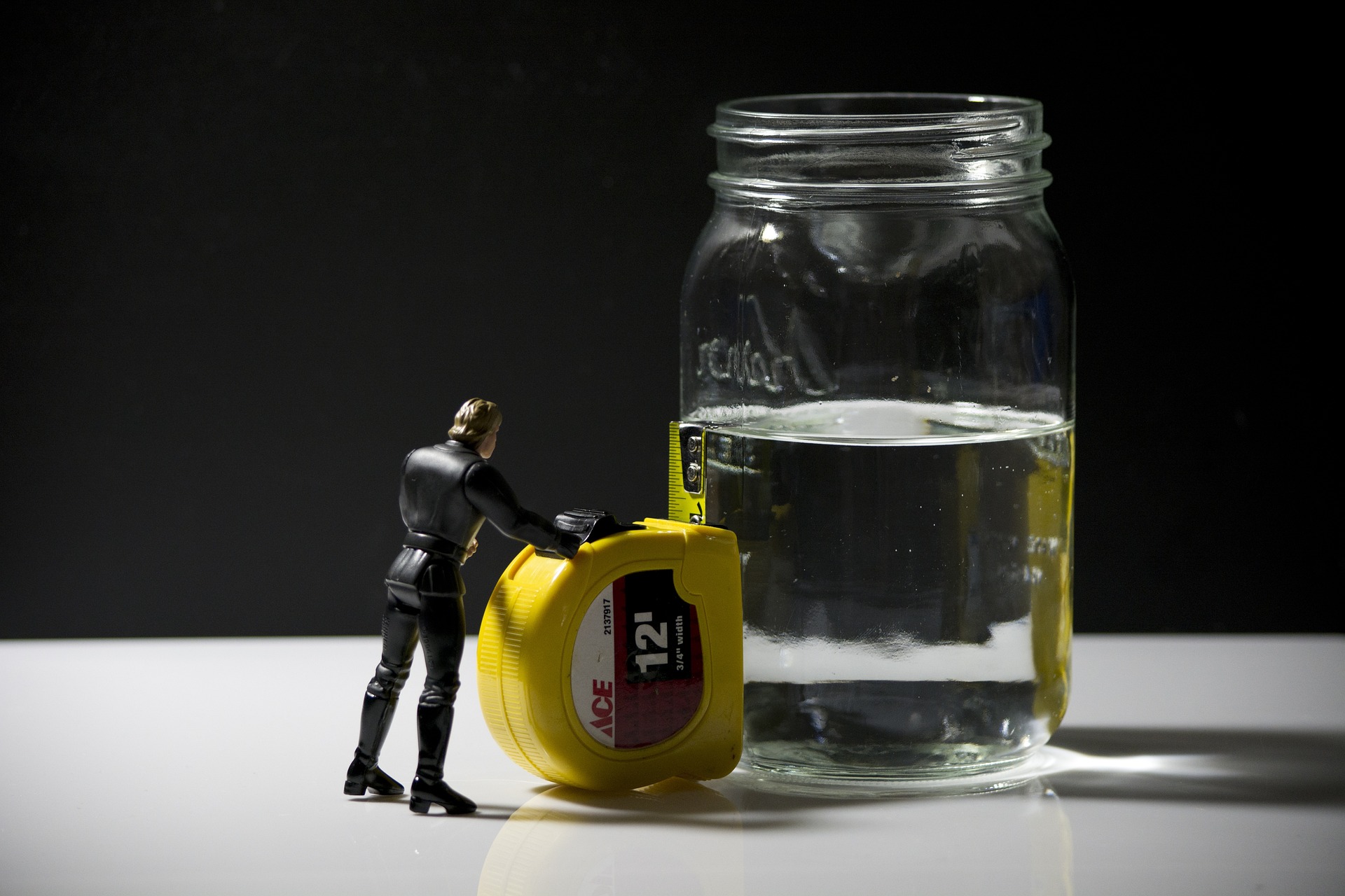 A toy figure assessing the level of water in a mason jar using a tape measurer.