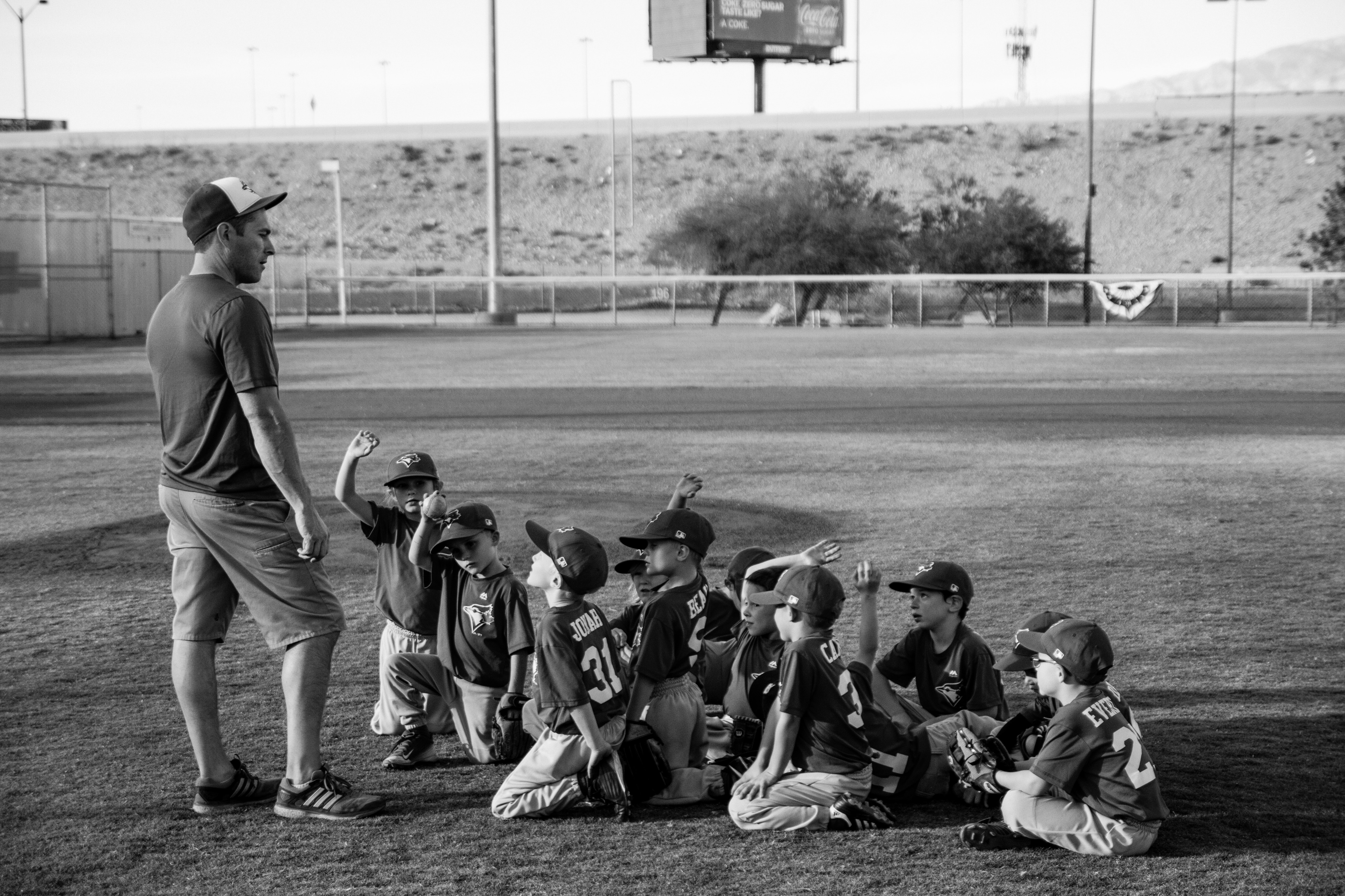 A coach talking to a team of very enthusiastic young kids, many with their hands raised.