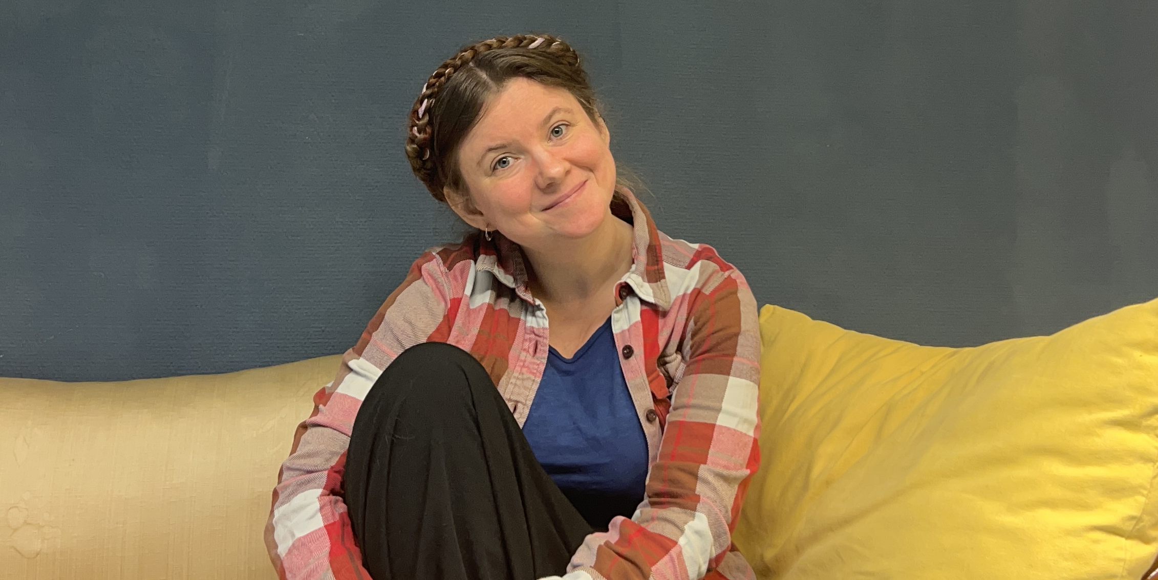 EsterDaniel sitting in a yellow sofa with their legs pulled up, in a long black skirt, red checked flanell shirt, braided hair, and a big smile. The background is blue.